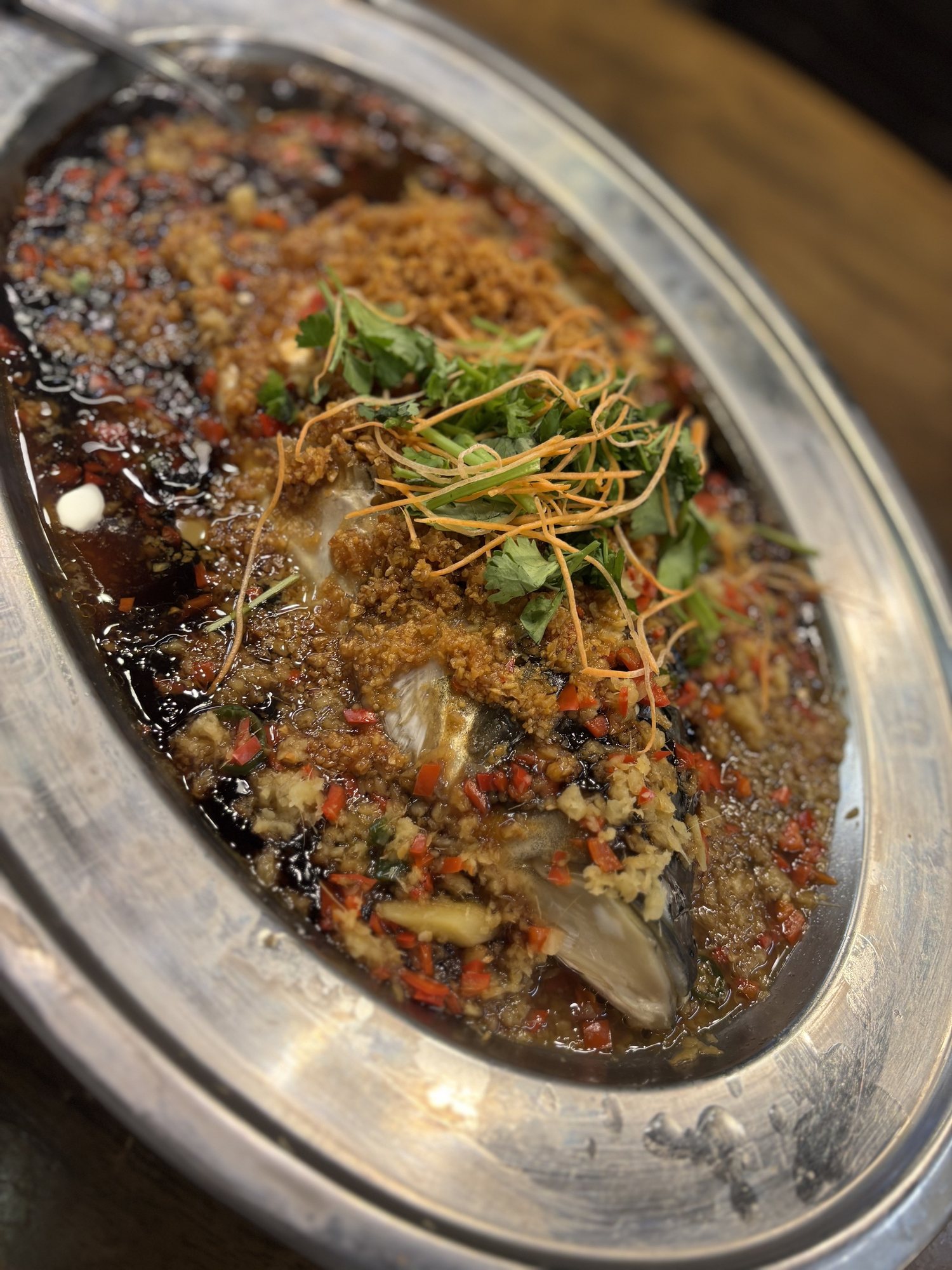 銀色の楕円の皿に蒸したナマズ。揚げにんにく、刻んだ赤い唐辛子、香菜、醤油色のタレ