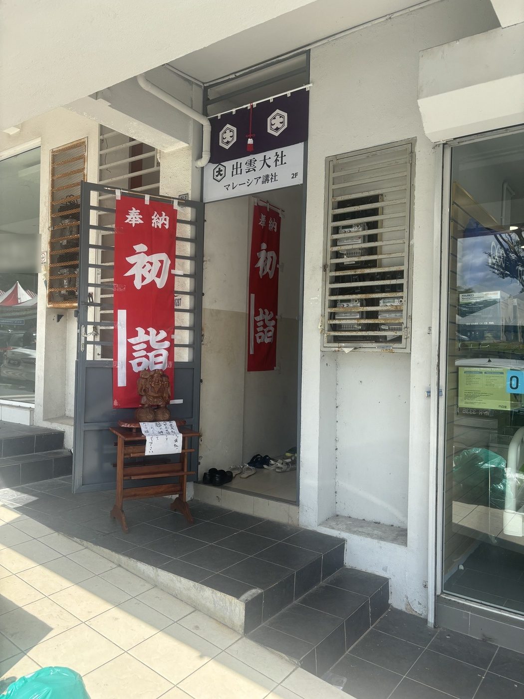 看板「出雲大社 マレーシア講社 2F」と赤い幟「奉納 初詣」、木彫りの大黒さま、入口に並ぶ靴